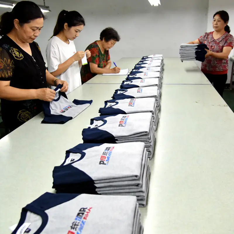 AQL 2.5 inspection — color, seams, sizing, print alignment Quality control inspector checking a finished sublimated esports jersey on a white inspection table with rulers, color-control charts, and a finished sample pinned to the wall in the background